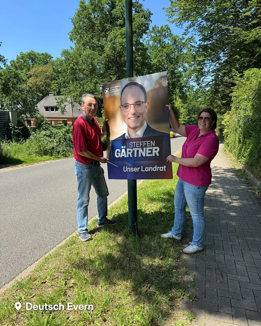 Zwei ehrenamtliche Helfer halten ein frisch montiertes Wahlplakat von Steffen Gärtner mit der Aufschrift „Unser Landrat" an einem Laternenmast in Deutsch Evern.